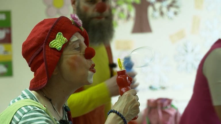 Eine Frau als Clown verkleidet mit roter Nase und roter Mütze bläst eine Seifenblase.