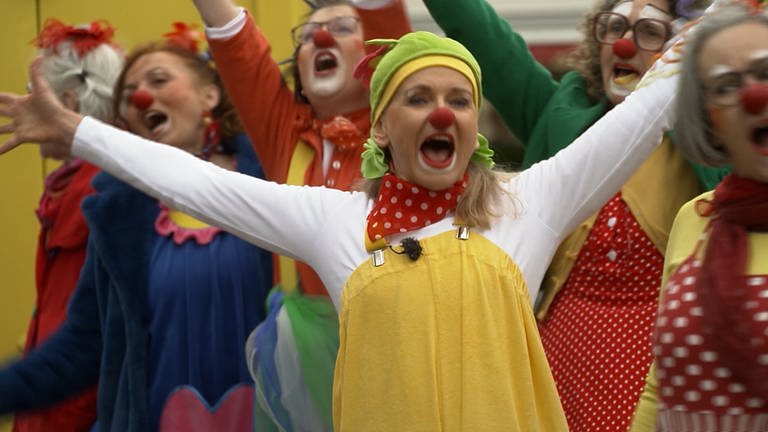 Eine blonde Frau mit gelb-weißem Clownsanzug, einer roten Nase und grün-gelber Mütze steht vor eine Gruppe Clowns.