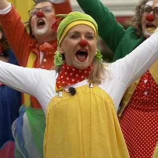 Eine blonde Frau mit gelb-weißem Clownsanzug, einer roten Nase und grün-gelber Mütze steht vor eine Gruppe Clowns.