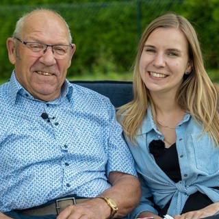 Ein älterer Herr und eine junge Frau sitzen im Freien auf einer gepolsterten Bank. Er trägt ein blaues Hemd und sie eine blaue Bluse.