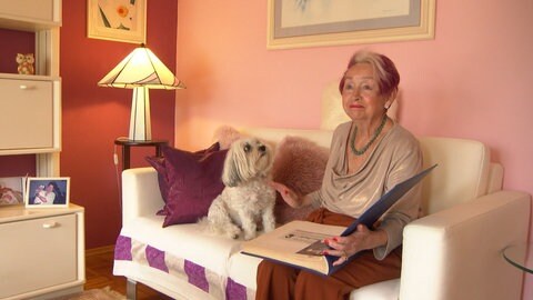 Brigitte sitzt rechts auf einem Sofa, neben ihr ein kleiner Hund. Sie hält ein Fotoalbum in der Hand.