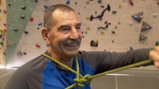 Der 89-jährige Franz steht in Sportkleidung vor einer Kletterwand in einer Kletterhalle und lächelt.