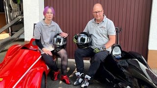 Jule und ihr Papa Andreas fahren leidenschaftlich gerne mit ihren Seifenkisten. Sie sitzen auf ihren Seifenkisten vor ihrer Garage. Beide haben einen Helm in der Hand und schauen in die Kamera. 