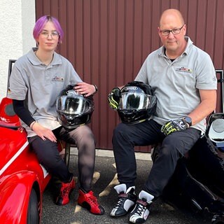 Jule und ihr Papa Andreas fahren leidenschaftlich gerne mit ihren Seifenkisten. Sie sitzen auf ihren Seifenkisten vor ihrer Garage. Beide haben einen Helm in der Hand und schauen in die Kamera. 