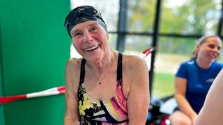 Die 83-jährige Margarete sitzt in einem schwarzen Badeanzug mit gelben und rosa Details auf einer Bank im Schwimmbad. Sie lacht in die Kamera. 