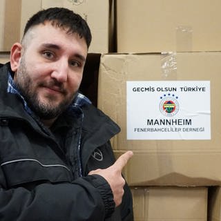 Mann vor einer Paketwand. Auf einem Paket klebt ein Zettel mit der Aufschrift: ‘Gute Besserung, Türkei!’