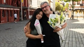 Ein Mann mit weißen Haaren und Brille und eine junge Frau mit schwarzen Haaren stehen auf einem Marktplatz.