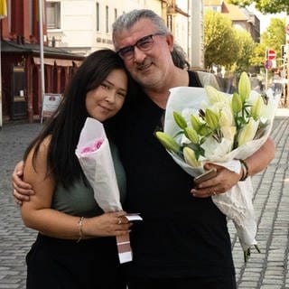 Ein Mann mit weißen Haaren und Brille und eine junge Frau mit schwarzen Haaren stehen auf einem Marktplatz.