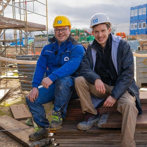 Zwei junge Männer sitzen in Arbeitskleidung auf einer Baustelle und lächeln in die Kamera.