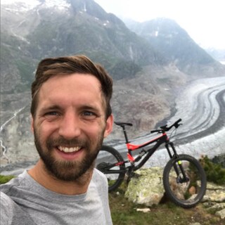 Junger Mann mit braunen Haaren lächelt in die Kamera, im Hintergrund Berge und ein Fahrrad