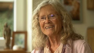 Frau mit langen grauen Haaren und Brille lacht.