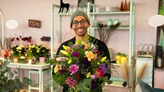 Mann steht in einem Blumenladen, hält einen bunten, großen Blumenstrauß und lächelt in die Kamera.