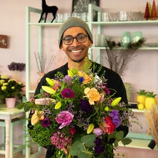 Mann steht in einem Blumenladen, hält einen bunten, großen Blumenstrauß und lächelt in die Kamera.