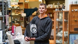 Dimitri hält eine Musikbox in der Hand und lächelt. Im Hintergrund erkennt man das Sozialkaufhaus, in dem viele gebrauchte Dinge für wenig Geld weiterverkauft werden. 