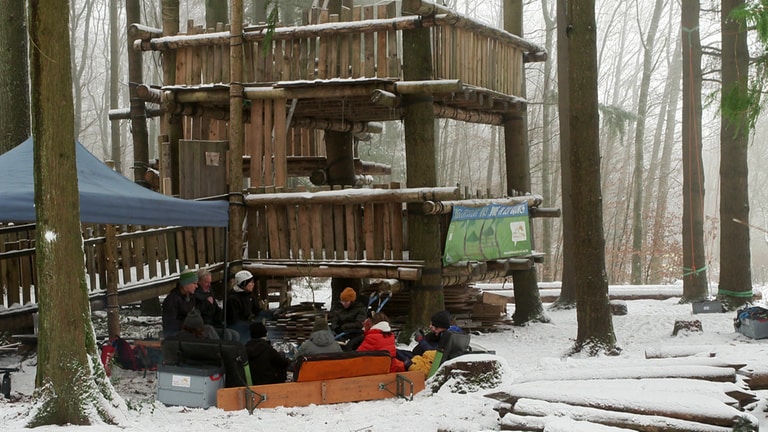 Im Vordergrund eine Gruppe junger Menschen, dick eingepackt, an einem Lagerfeuer. Im Hintergrund ein Wald mit einem mehrstöckigen Baumhaus. 