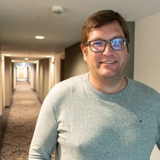 Hotelinhaber Alexander Wurster steht in einem langen, leeren Flur und blick in die Kamera