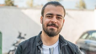 Mahmoud, junger Mann, Mitte 20, Vollbart mit nach hinten gegelten langen Haaren, einer schwarzen Jacke in einem Hof stehend.