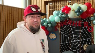 Ein Mann mit Spiderman-Kappe steht vor einer Sprossenwand mit Ballons und Superheldendekoration.