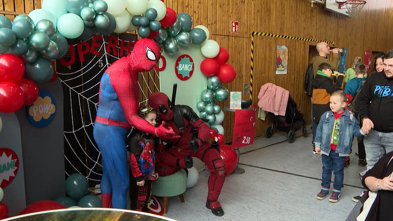 Ein Mann im Spiderman-Kostüm und ein Mann im Deadpool-Kostüm posieren mit einem kleinen Kind für ein Foto.