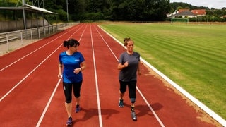 Zwei Frauen joggen auf einem Sportplatz. Die rechte Frau hat eine Beinprothese