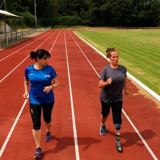 Zwei Frauen joggen auf einem Sportplatz. Die rechte Frau hat eine Beinprothese
