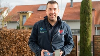 Ein Mann mittleren Alters und in blauer Uniform der Mobile Retter steht im Garten und hält ein Handy und ein kleines Mäppchen in der Hand.