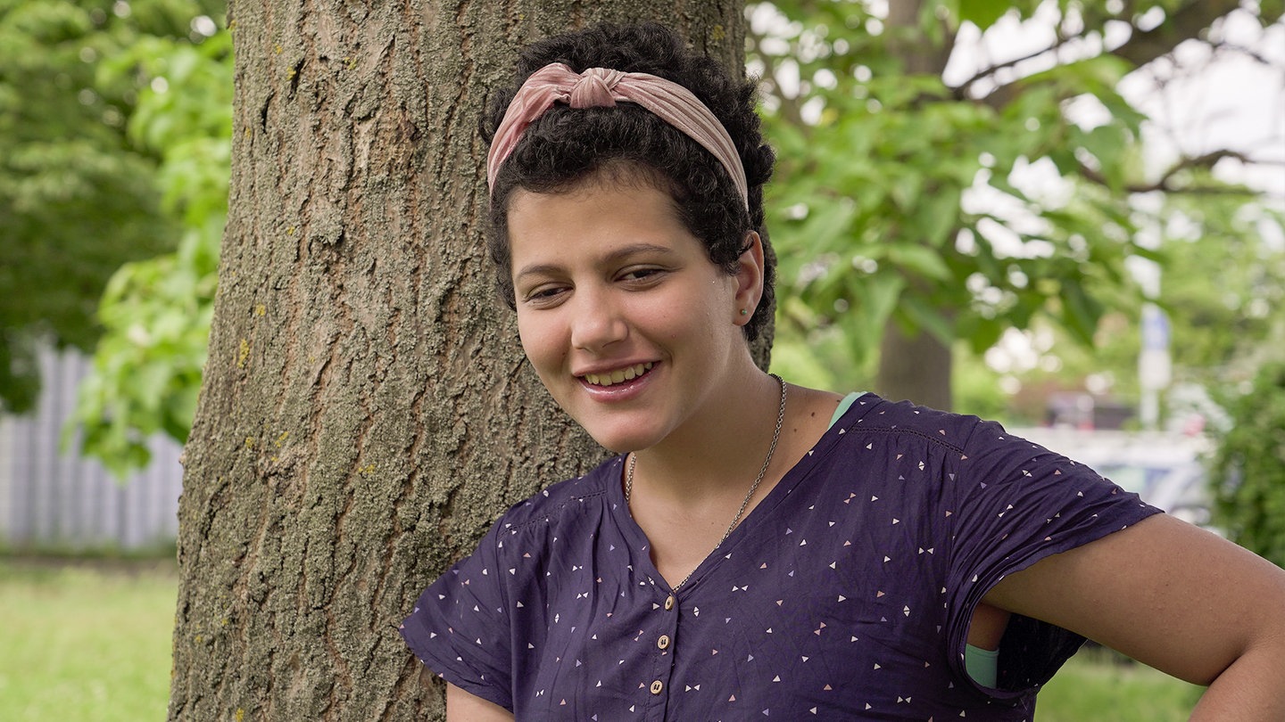 Maja, vor einem Baum. Sie hat ein Haarband in ihren kurzen, braunen Haaren