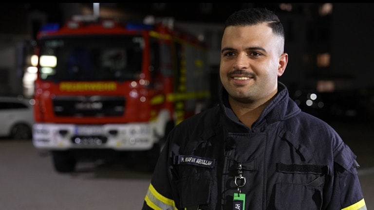 Ein Mann mit Feuerwehranzug steht in einer Feuerwehrwache und lächelt.