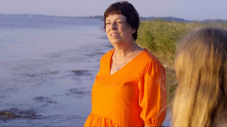 Eine ältere Frau mit kurzen braunen Haaren und orangefarbenen Kleid steht am Meer und schaut in die Sonne.