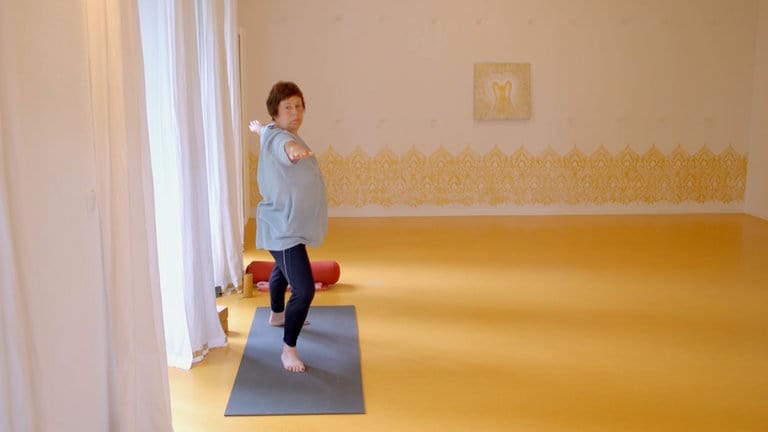 55-jährige Frau, die mit einem weißen T-Shirt, kurzen roten Haaren in einem Raum mit Holzboden. Sie ist in einer Yogaposition auf einer grauen Matte.