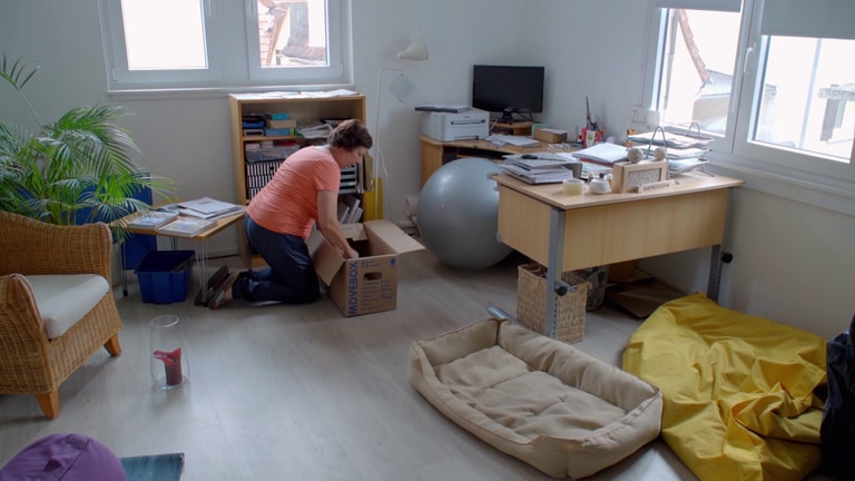 Eine ältere Frau in apricotfarbenen T-Shirt packt in einem Büro einen Karton.