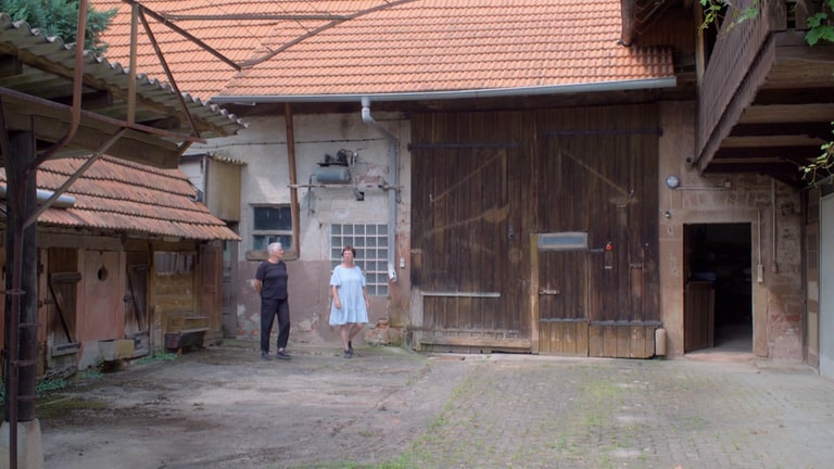 Zwei ältere Frauen in Sommerkleidung laufen über den Hof eines alten Bauernhofs.