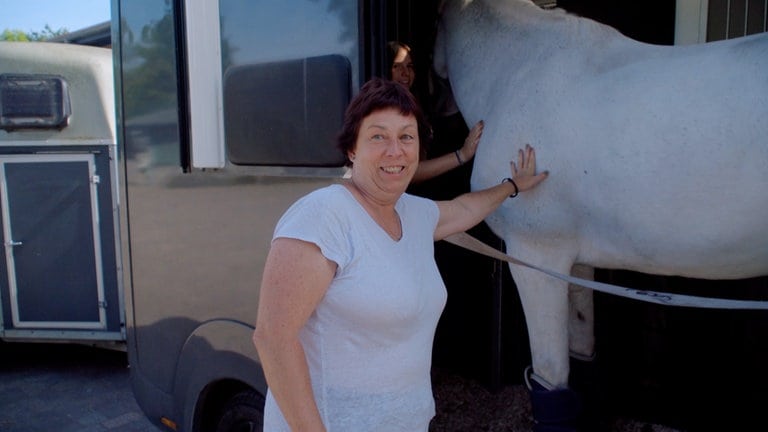 55-jährige Frau, die mit einem weißen T-Shirt, kurzen roten Haaren an der Seite eines weißen Pferdes steht und es berührt.