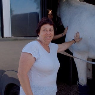 55-jährige Frau, die mit einem weißen T-Shirt, kurzen roten Haaren an der Seite eines weißen Pferdes steht und es berührt.