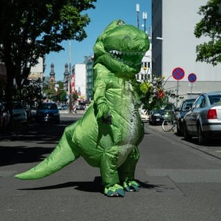 Ein Mann mittleren alters posiert mitten auf einer Straße in einem Dinokostüm mit Blumen in der Hand