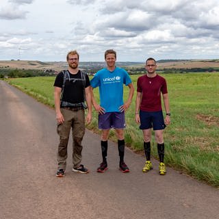 Drei junge Männer stehen in Wanderklamotte im Feld