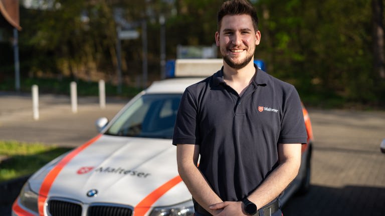 Junger Mann mit kurzen Haaren im Outfit der Malteser steht vor einem Malteser-Einsatzwagen. 