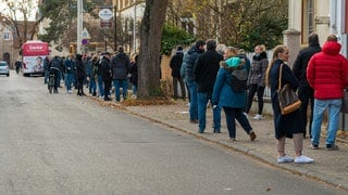 Menschen stehen in einer Reihe, ganz am Ende der Schlange sieht man den roten Impfbus.