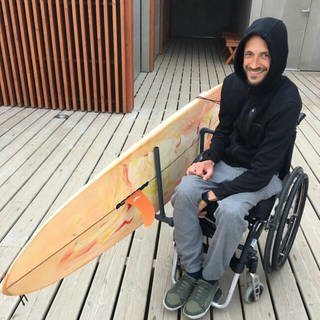 Der querschnittgelähmte Johannes Laing sitzt lächelnd in seinem Rollstuhl, an dem mit Hilfe einer selbstgebauten Halterung sein Surfbrett befestigt ist.