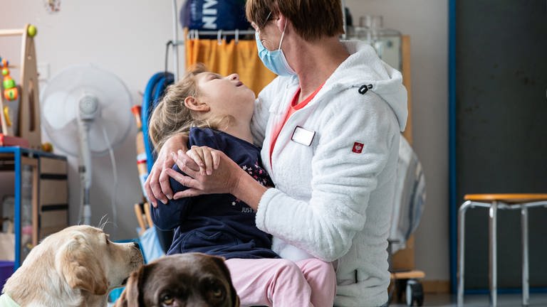 Palliativschwester Ivana hält ein krankes Mädchen im Arm, vorne liegen zwei Hunde.