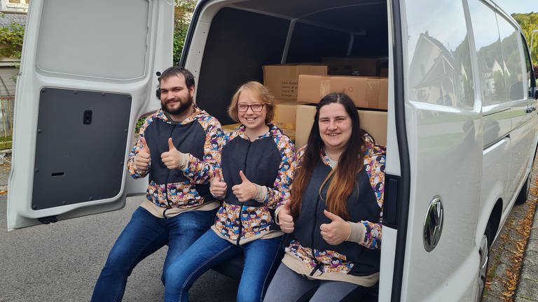 Drei junge Menschen sitzen im Kofferraum eines Kastenwagens, der mit vielen Kartons geladen ist.