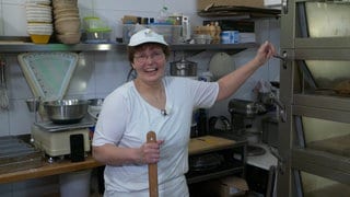 Frau mit Brille, kurzen Haaren und weißer Mütze und T-Shirt steht am Ofen in der Bäckerei