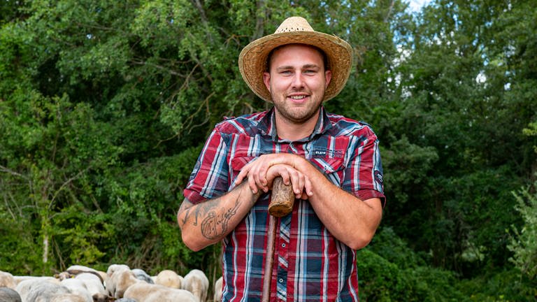 Junger Mann mit kariertem Hemd und Strohhut lehnt vor einem Wald auf einem Stock. Um ihm herum sind Schafe. 