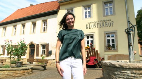 Junge Frau mit braunen Haaren und grünem T-Shirt steht lächelnd vor Gebäude.