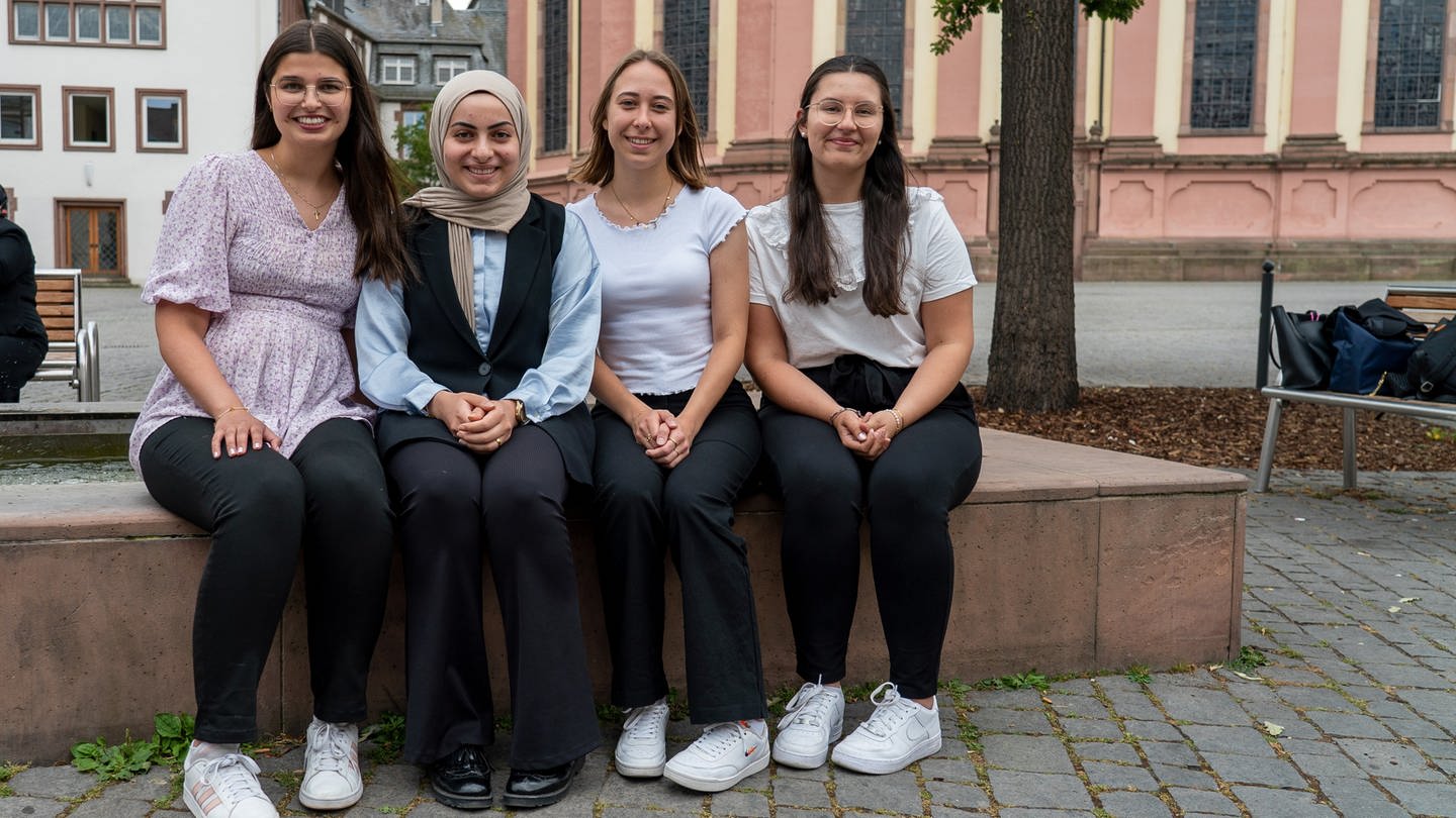 Vier junge Finanzbeamtinnen sitzen nebeneinander auf dem Rand eines Brunnens auf dem Wormser Marktplatz und lächeln.