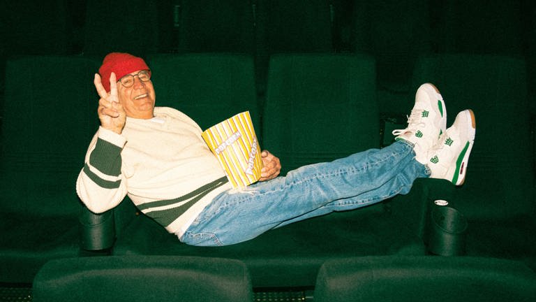 Ältere Person in Street-Style Bekleidung, liegt quer auf Sesseln in einem Kinosaal. Hält dabei eine Tüte Popcorn und macht das Peace Zeichen.
