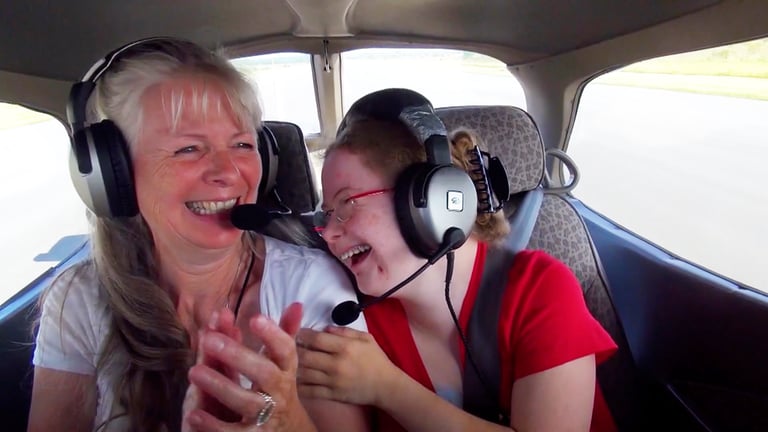 Eine junge und eine ältere Frau mit Kopfhörern sitzen in einem Flugzeug und lachen. 