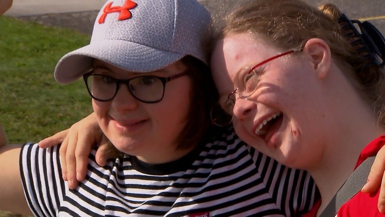 Zwei junge Frauen mit Brille lächeln und lehnen Arm in Arm aneinander.