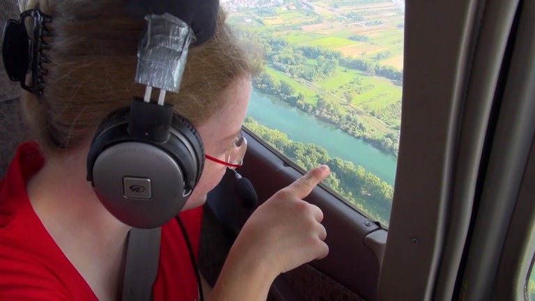 Junge Frau schaut aus einem Flugzeugfenster ins Rheintal und zeigt nach draußen.