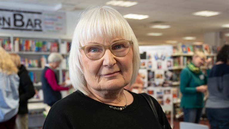 Ältere Dame, mit langen, glatten, weißen Haaren und Brille. Steht in einer Bücherei und schaut in die Kamera. 